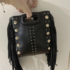Maje väska - Cool svart axelväska från Maje i skinn med fransar på sidorna och guldiga nitar framtill. Väskan har ett handtag upptill och avtagbar axelrem. Den är i Lilla storleken 
