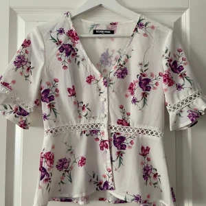 Vit blommig blus från Fashion Union - Söt vit blus med blommigt mönster i lila och rosa toner från Fashion Union. Blusen har v-ringning, knappar framtill, volangdetaljer på ärmarna och spetsdetalj vid midjan. Perfekt för vår och sommar!