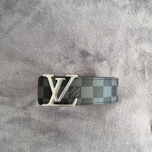 Lv bälte  - Snyggt bälte från Louis Vuitton med klassiskt rutigt mönster i svart och grått. Bältet har ett stort LV-spänne i silver och insida i ljust skinn. Perfekt accessoar för att lyfta din outfit.