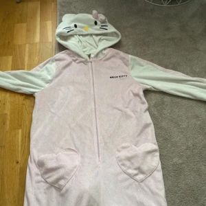 Hello Kitty jumpsuit med luva - Supergullig ljusrosa och vit Hello Kitty jumpsuit med dragkedja och huva med öron och ansikte. Två stora hjärtformade fickor framtill. Perfekt för dig som älskar söta och mysiga plagg!