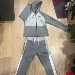 Grå tracksuit från Nike med vita detaljer - Säljer en grå tracksuit från Nike med vita sidor och dragsko i midjan. Byxorna har en sportig look och är perfekta för chill eller träning. Klassisk Nike-logga på benet och elastisk midja för skön passform.