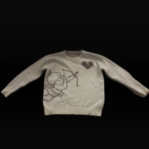 Vit stickad tröja med cupido-tryck - Vit stickad tröja med motiv av en cupido som skjuter pil mot ett hjärta på framsidan. På baksidan står texten 'I WOULD RATHER NOT LOVE' och BMC. Tröjan har långa ärmar och rund hals.