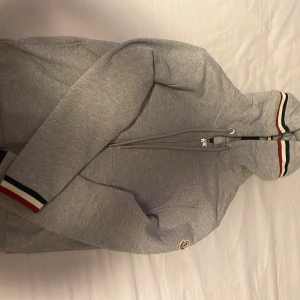 Grå hoodiejacka med dragkedja från Moncler - Grå hoodiejacka från Moncler med dragkedja framtill och huva med dragsko. Ränder i rött, vitt och blått på ärmslut och huva. Klassisk logotyp på bröstet. Perfekt för en sportig och stilren look.