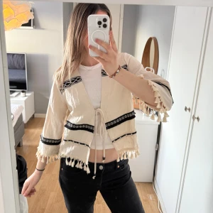 Beige blus med svarta detaljer från Zara - Säljer en beige blus/kofta från Zara med svarta broderade band och fransar vid ärmslut och nederkant. Jackan har knytning framtill och trekvartsärmar. Superfint skick!💕.