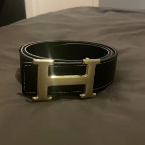Svart bälte med guldfärgat H-spänne - Snyggt svart bälte med ett stort H-format spänne i guld. Bältet har vita sömmar längs kanterna och är gjort i skinn. Perfekt accessoar för att lyfta din outfit. 105