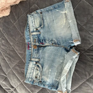 Ljusblå jeansshorts med fransar från Replay - Säljer ett par ljusblå jeansshorts från Replay med slitna detaljer och fransig kant. Shortsen har klassiska fickor fram och bak, samt synliga sömmar och knappstängning. Perfekta för sommaren!