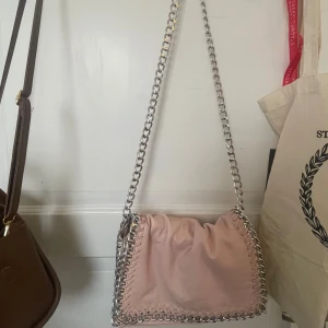 Ljusrosa axelväska med kedjedetaljer från Tiamo - Säljer en ljusrosa axelväska från Tiamo med silverfärgad kedja som axelrem och runt kanterna. Liknar Stella McCartney väskorna! Den är väldigt sliten så därför säljer jag den billigt. 