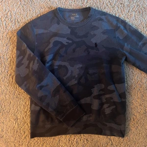Blå camouflagemönstrad tröja från Polo Ralph Lauren - Säljer en blå långärmad tröja från Polo Ralph Lauren med camouflagemönster. Tröjan har rund halsringning och en liten broderad logga på bröstet. Perfekt för dig som gillar streetwear och vill ha något unikt i garderoben. Står storlek M men passar mer som en S