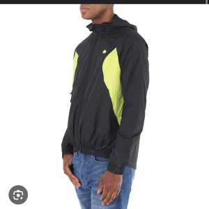Svart och gul vindjacka från Lacoste - Säljer en snygg svart vindjacka från Lacoste med gula detaljer på sidorna och bröstet. Jackan har huva, dragkedja och resår i midjan. En skön windbreaker i bra skick.