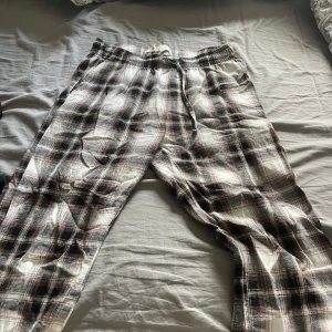 Rutiga mjukisbyxor med snörning - Säljer ett par rutiga mjukisbyxor i svart, vitt och brunt med elastisk midja och snörning. Perfekta för en avslappnad stil och riktigt sköna att ha på sig hemma eller till vardags.