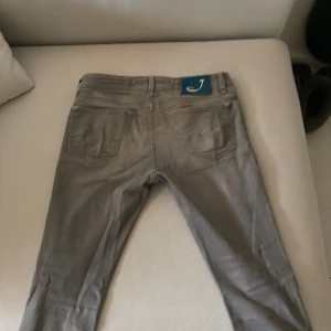 Grå jeans från Jacob Cohën - Säljer ett par grå jeans från Jacob Cohën med klassisk femficksdesign och silverfärgad knapp. Byxorna har en normal passform och är tillverkade av exklusiva material med italienskt hantverk. Perfekta för dig som gillar stilrena och snygga jeans. Däremot finns defekter, skriv för bilder.