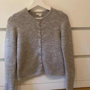 Ljusgrå stickad kofta från Gina Tricot - Mysig långärmad ljusgrå stickad kofta från Gina Tricot med knappar framtill. Den är från Gina Young i storlek 146/152 och är i bra skick, har endast använt några gånger. Säljer eftersom den är för liten. Lite lite nopprig. 
