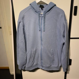 Ljusblå hoodie med dragkedja från H&M - Mysig ljusblå hoodie från H&M i relaxed fit. Tröjan har dragkedja framtill, två fickor och huva med dragsko. Perfekt för en avslappnad stil och enkel att matcha med jeans eller joggers.