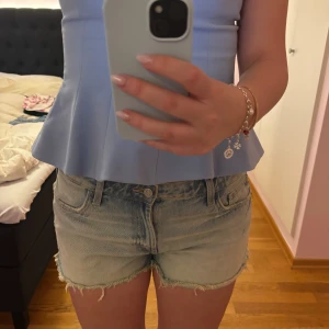 Ljusblå jeansshorts med fransar - Säljer ett par ljusblå jeansshorts med råa franskantade benslut. Klassisk femficksmodell med knapp och dragkedja framtill. Perfekta för varma dagar!