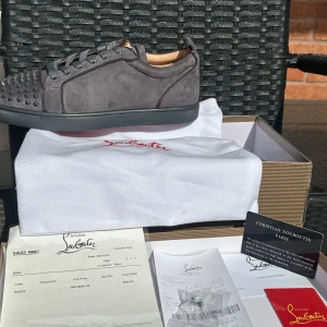 Gråa sneakers med nitar från Christian Louboutin - Säljer ett par gråa sneakers från Christian Louboutin i mocka med coola nitar framtill och klassisk röd sula. Skorna har snörning och rund tå, perfekt för dig som vill sticka ut med din stil.