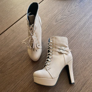 Beige höga boots med snörning och spännen - Säljer ett par beige boots med höga klackar och platåsula. Skorna har snörning framtill, två dekorativa spännen på sidan och rund tå. Tillverkade i syntetmaterial och passar perfekt för dig som vill sticka ut.
