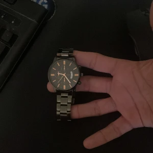 Svart och silverfärgad herrklocka från curren - Stilren herrklocka från Curren med svart urtavla och orange detaljer. Klockan har ett robust silverfärgat metallarmband. Perfekt accessoar för dig som gillar klassisk design. behöver batteri 