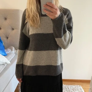 Randig stickad tröja från Vero Moda - Mysig stickad tröja från Vero Moda med breda grå och beige ränder. Tröjan har lång ärm och en avslappnad passform, perfekt för kyligare dagar. Klassisk rund halsringning och mjukt material.