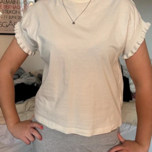 Vit t-shirt med volangärm från Zara - Säljer en söt vit t-shirt från Zara med volangdetaljer vid ärmsluten. T-shirten har en rak passform och är kortärmad, perfekt för en enkel och stilren look.