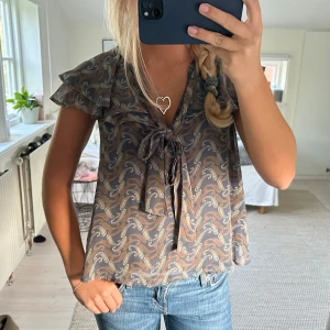 Mönstrad blus med volangärm och knyt - Säljer en luftig blus  med korta volangärmar och knyt framtill vid halsen. Perfekt till jeans eller kjol för en snygg look.