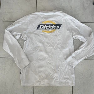 Vit långärmad tröja från Dickies - Vit långärmad tröja från Dickies med tryckt logga i gult och svart både fram och bak. Klassisk rund halsringning och rak passform. Perfekt för en avslappnad stil. Passar både S och M. Fläck på vänster arm (se bild 4) därav billigare.