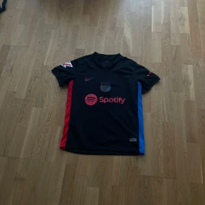 FC Barcelona svart bortatröja Nike - Säljer en svart FC Barcelona bortatröja från Nike med röda och blå sidopaneler. Tröjan har korta ärmar, Spotify-logga på bröstet och UNHCR-tryck på ryggen. Perfekt för dig som vill visa ditt stöd för Barca!Storlek M i barn
