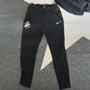 Svarta Nike Dri-Fit AIK träningsbyxor - Svarta träningsbyxor från Nike med AIK-logga på ena benet och Nike Swoosh på andra. Byxorna har elastisk midja och är tillverkade i Dri-Fit-material som andas. Perfekta för fotbollsträning eller sportiga dagar.