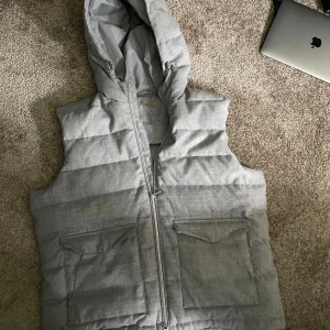 Grå dunväst med huva från Stenströms - Snygg grå dunväst från Stenströms med huva och dragkedja framtill. Västen har två stora fickor med lock och en quiltad design som ger en modern look. Perfekt för sommaren