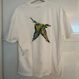 Vit t-shirt med fågeltryck från Axel Arigato - Vit T-shirt med tryck på ryggen.  Strl L. Normal i strl och passar både dam och herr.  Chest 53 Length 65 Är i ny skick.  Nypris: 1250kr