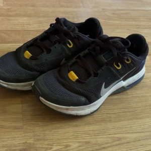 Nike Air Max sneakers i svart och gult - Säljer ett par svarta Nike Air Max sneakers med gula detaljer och blå AIR-tryck på hälen. Skorna har snörning, vit sula och är tillverkade i syntetmaterial med mesh för bra ventilation. Perfekta för dig som gillar sportig stil.