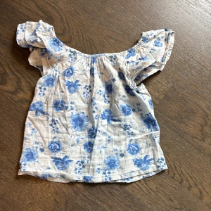 Blommig blus med volangärmar från Lindex - Söt blus från Lindex i vitt med blått blommönster. Toppen har korta volangärmar och smockad rygg för extra komfort. Perfekt för varma dagar och enkel att matcha med jeans eller kjol.