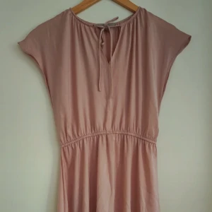 Ljusrosa klänning - Säljer en fin ljusrosa klänning. Klänningen har korta ärmar, knytning vid halsen och resår i midjan. Den är tillverkad i ett mjukt och lätt material och har en avslappnad passform.