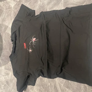 Svart t-shirt med röd grafisk print och spindelmotiv - Cool svart t-shirt från Off-White med stor röd grafisk print och spindelmotiv på ryggen samt mindre tryck framtill. Klassisk passform och korta ärmar. Perfekt för dig som gillar streetwear och unika detaljer.