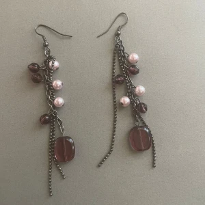Långa örhängen med lila pärlor och kedjor - Säljer ett par långa örhängen med mörklila och ljusrosa pärlor samt dekorativa kedjor. Örhängena har en design med flera hängen och stora lila glaspärlor längst ner. Perfekta för att ge din stil ett lyft!