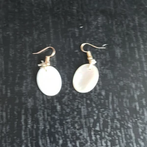 Vita ovala örhängen med pärldetalj - Säljer ett par eleganta örhängen med ovala vita hängen och små pärldetaljer. Krokarna är i guldfärg och örhängena har en stilren och klassisk look som passar till många tillfällen.