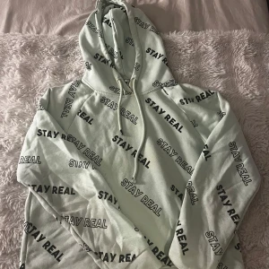 Ljus grön hoodie  - Säljer en hoodie med svart text 'STAY REAL' tryckt över hela tröjan. Tröjan har huva med dragsko och långa ärmar. Perfekt för en avslappnad och cool stil.