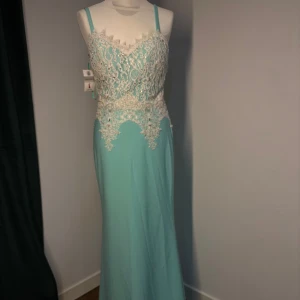 Ljusblå långklänning med spetsdetaljer - Ljusblå långklänning med smala axelband och vacker vit spets upptill. Klänningen har figurnära passform och spetsen är dekorerad med pärlor och broderier både fram och bak. Perfekt för bal eller fest.  Helt ny från företag.