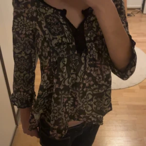 Jättesöt Odd Molly blus 🪻 - Supersnygg blus från Odd Molly i svart med grått, rosa och grönt mönster. Blusen har v-ringning med svarta spetsdetaljer och en söt rosett framtill. Den har trekvartsärmar med lätt volang. Perfekt för dig som gillar unika plagg. I storlek 1 i odd Molly vilket motsvarar i första hand xs men även s. Lätt figursydd i tunt tyg.🪻