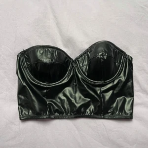 Svart glansig bustier från Monica Geuze - NAKD - Säljer en svart glansig bustier från Monica Geuze/ NAKD. Toppen är axelbandslös med markerade kupor och dragkedja bak. Perfekt att styla med kavaj eller på egen hand för en trendig look.