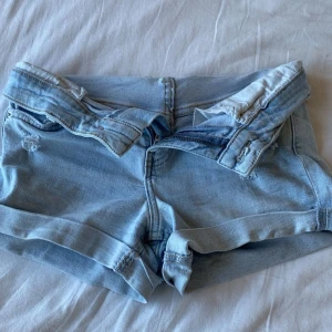 Ljusblå jeansshorts från Bershka - Säljer ett par ljusblå jeansshorts från Bershka med upprullade benslut och slitna detaljer framtill och baktill. Klassisk femficksmodell med knapp och dragkedja. Perfekta för varma dagar. Högmidja men jättafina att vika ner till lägre midja!