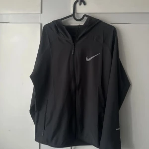 Svart vindjacka med huva från Nike - Säljer en svart vindjacka från Nike med huva och dragkedja framtill. Jackan har långa ärmar och två sidofickor med dragkedja. Perfekt för utomhusaktiviteter och träning. Swoosh-logga på bröstet.