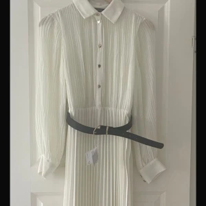 Vit plisserad skjortklänning med bälte - Säljer en elegant vit plisserad skjortklänning med klassisk krage och knappar framtill. Klänningen har långa ärmar och ett svart bälte i midjan som ger en snygg siluett. Perfekt för dig som gillar stilrena och tidlösa plagg.