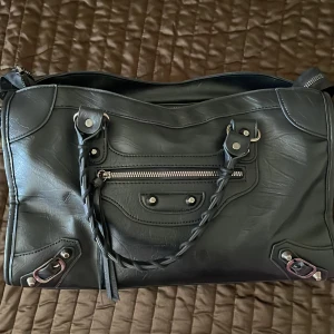 Svart handväska med flätade handtag - Säljer en svart handväska med snygga flätade handtag och silverfärgade metalldetaljer. Väskan har en dragkedja framtill och dekorativa nitar. Perfekt för dig som vill ha en stilren och rymlig accessoar till vardagen.