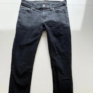 Svarta jeansbyxor från Nudie Jeans - Säljer ett par svarta jeansbyxor från Nudie Jeans med klassisk femficksdesign och raka ben. Jeansen har normal passform och är tillverkade i denim med snygga detaljer vid fickorna. Perfekta för dig som gillar stilrena och tidlösa jeans.