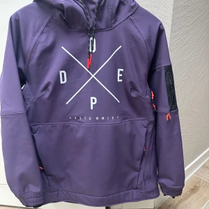 Lila jacka  med huva från Dope S - Snygg lila jacka från Dope med stor huva och tryck framtill. Jackan har dragkedjor i rött på fickorna och en extra ficka på ärmen. Perfekt för utomhusaktiviteter 
