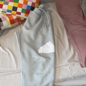 Ljusblå mjukisbyxor från Adidas - Säljer ett par ljusblå mjukisbyxor från Adidas med resår i midjan och dragsko. Byxorna har en vit Adidas-logga på benet och är perfekta för en avslappnad stil. Sköna och stilrena byxor med normal passform.
