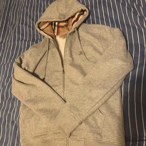 Grå hoodie med dragkedja från Burberry - Säljer en grå hoodie med dragkedja från Burberry. Jackan har klassiskt rutigt foder i huvan och insidan, samt snörning i huvan. Perfekt för dig som gillar stilrena och bekväma plagg med en exklusiv touch.