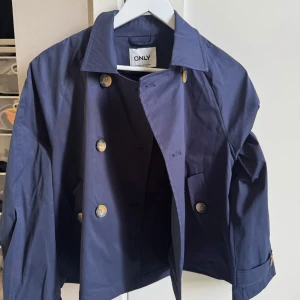 Mörkblå trenchcoat från ONLY - Säljer en mörkblå kappa från ONLY med dubbelknäppning och klassisk krage. Jackan har guldiga knappar och två fickor framtill. Perfekt för dig som vill ha en stilren och tidlös look.