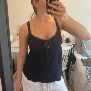 Mörkblå topp med spetsdetaljer - Säljer en mörkblå topp med spetsdetaljer vid axelbanden och en knytning framtill. Toppen har en texturerad yta.