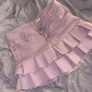 Ljuslila volangkjol i jeans - Superfin ljuslila jeanskjol med volanger och hjärtformade nitar. Kjolen har klassiska jeansfickor fram och bak samt dekorativ söm på bakfickorna. Perfekt för dig som gillar en söt och trendig stil. har sytt själv, syns på sömmarna innuti men inget alls utifrån.